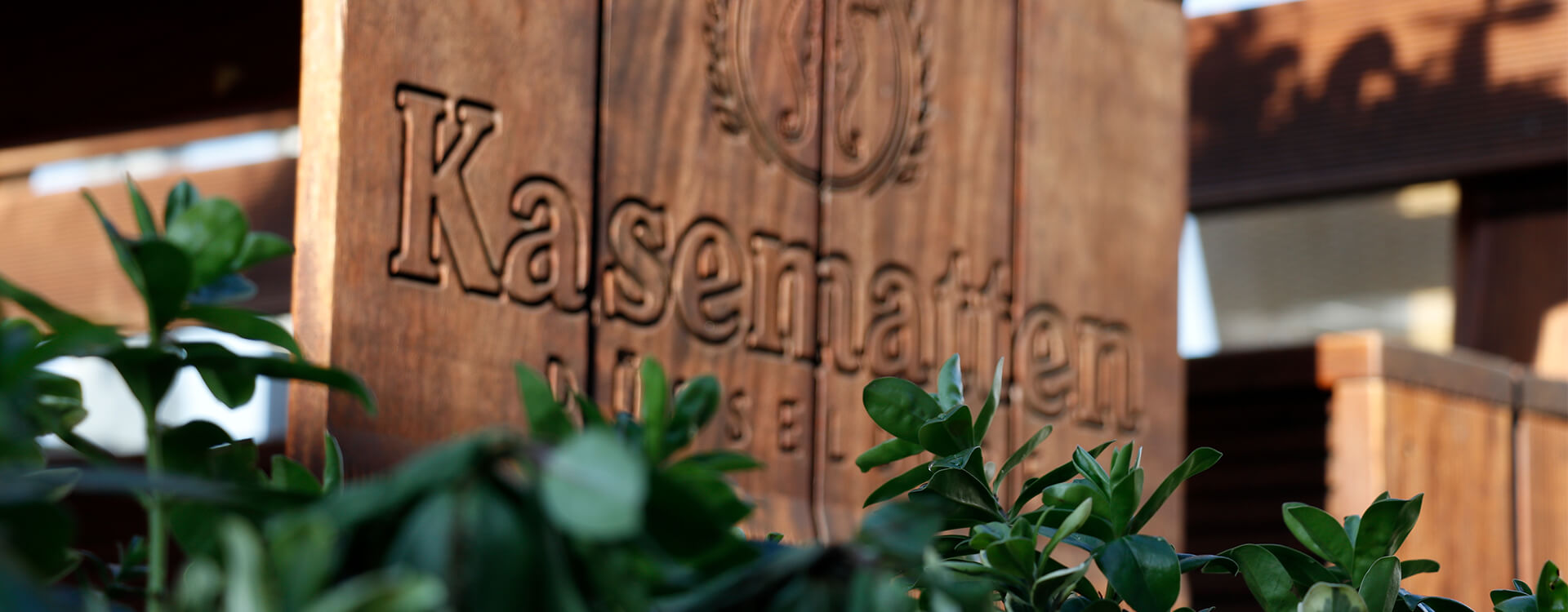 Ein Holzschild mit dem Logo der Kasematten eingraviert, steht vor einem Busch.